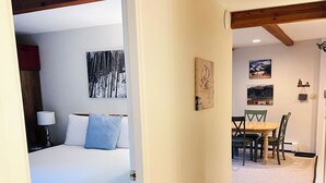 A bedroom with a bed, a nightstand, and a lamp. There is a dining area with a table and chairs. - Suite Situation in the Ski Village (Taos Ski Valley)