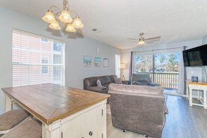 A spacious living area with a wooden dining table, two sofas, a ceiling fan, and large windows.