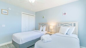 Quarto de hotel com duas camas, um ventilador de teto, uma porta branca e uma pequena mesa de cabeceira com abajur.