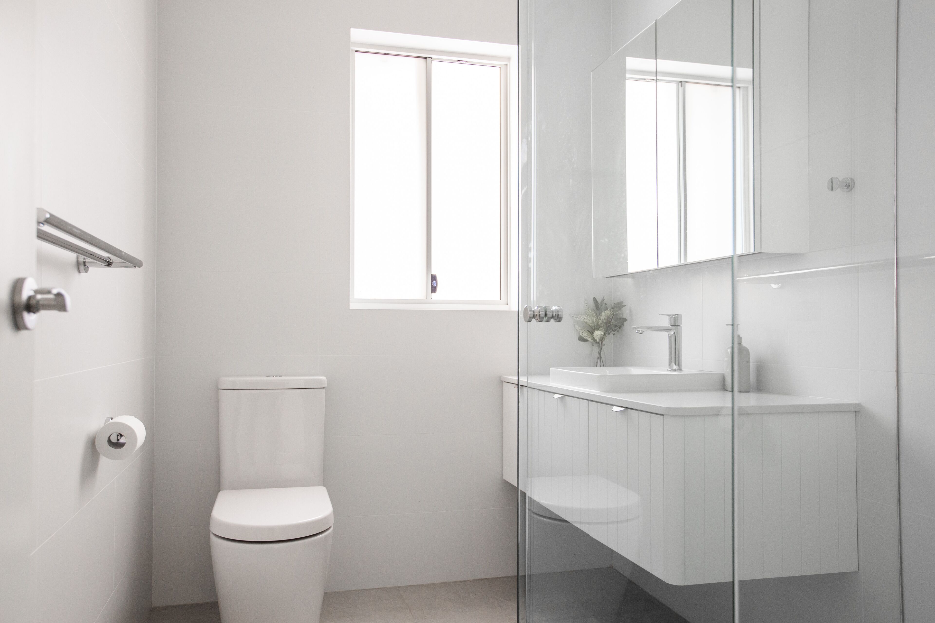 Et moderne badeværelse med toilet, glasvæg omkring brusenichen og et hvidt håndvaskarrangement med spejl.