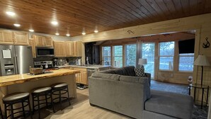 A modern kitchen with wooden cabinets, a central island with stools, a grey sofa, and a large window providing a view of the outdoors. - Cozy Lakefront Cottage on Little Pequaywan ~ Great for all seasons (Duluth)