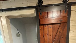A wooden barn-style sliding door with metal hardware, leading to a room with a white wall and a small hanging decoration. - Cozy Lakefront Cottage on Little Pequaywan ~ Great for all seasons (Duluth)