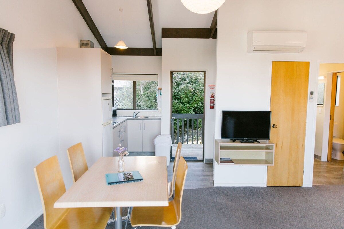 A modern room with a dining table, chairs, a television, and a kitchen area visible through an open door.