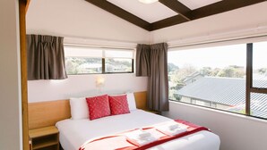 A hotel room with a large bed, a nightstand, a window with curtains, and a view of buildings outside. - Two Bedroom Apartment Sea View (Kaiteriteri)