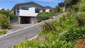 A two-story house with a driveway leading to a gated entrance, surrounded by lush greenery and a hilly landscape. - Two Bedroom Apartment Sea View (Kaiteriteri)
