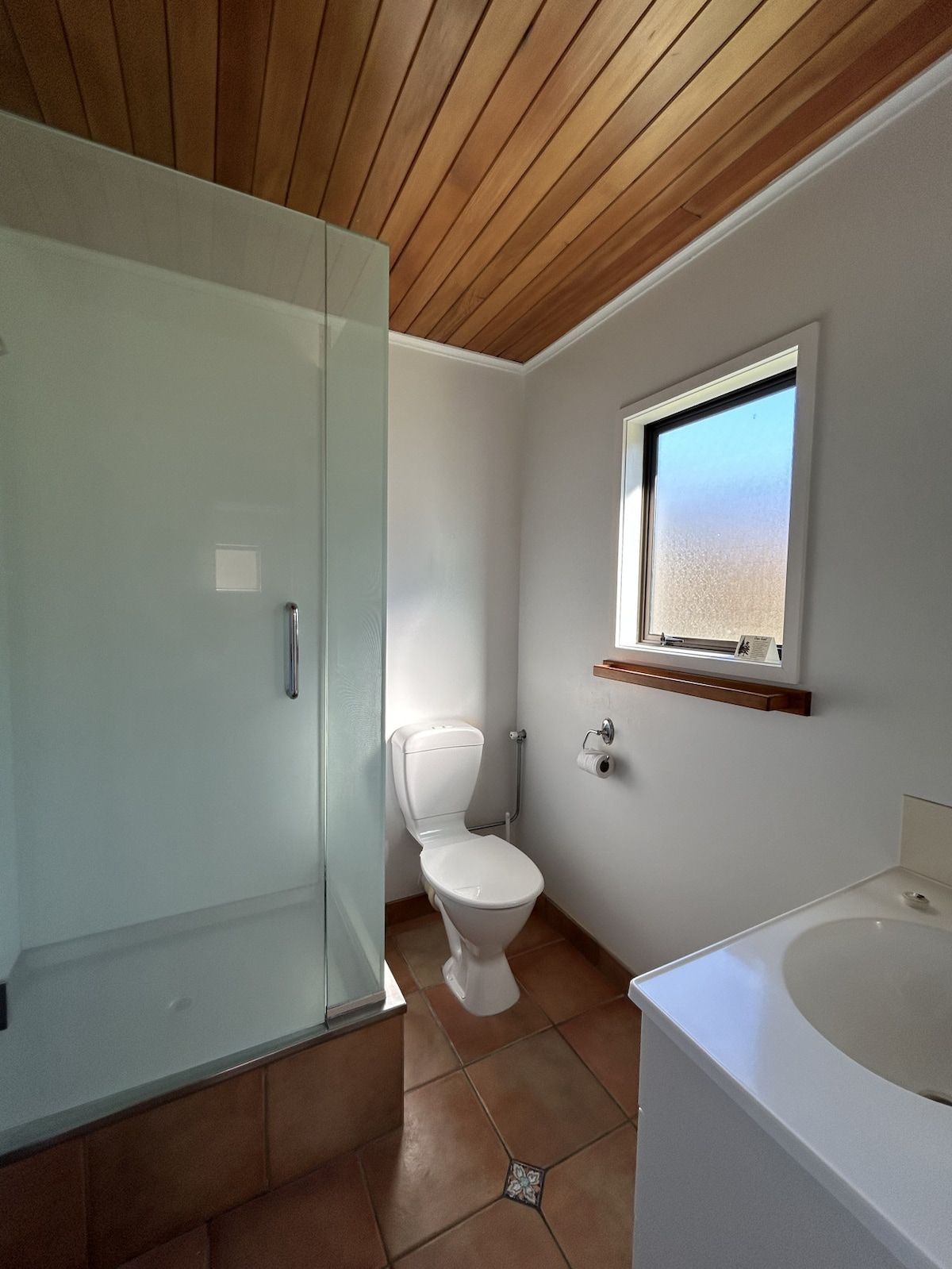 Une salle de bain avec une douche à parois de verre, des toilettes blanches, une fenêtre et un lavabo.