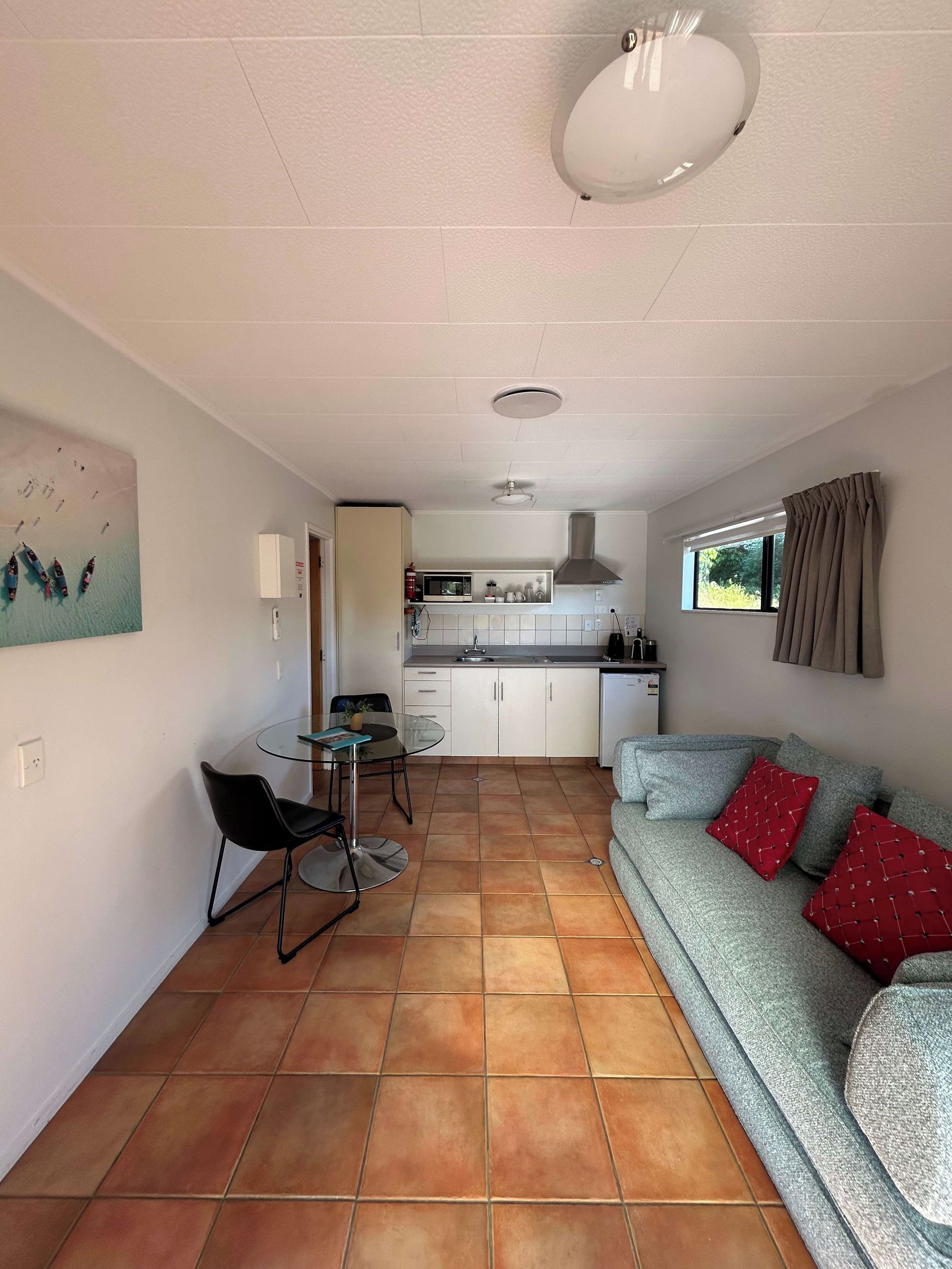 A compact living space with a kitchenette, a dining area with a glass table and chairs, and a sofa with red cushions.