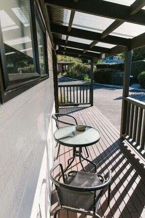 A covered outdoor seating area with a table and chairs, a view of the surrounding garden, and a building in the background.
