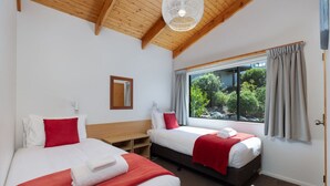 A bedroom with two beds, a wooden ceiling, a window with curtains, and a view of greenery outside. - Two Bedroom Garden View (Kaiteriteri)
