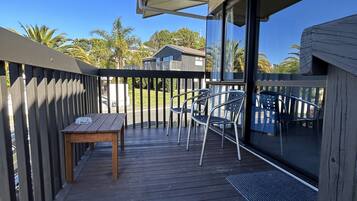 A wooden deck with a table and chairs, a glass door leading to a covered area, and a view of palm trees and a house in the background.