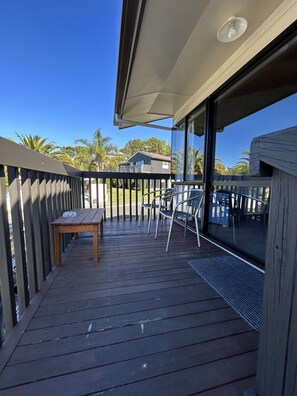 Una terraza de madera con una mesa y sillas, un ventanal de vidrio que da a un área cubierta, y una vista de palmeras y una casa al fondo.