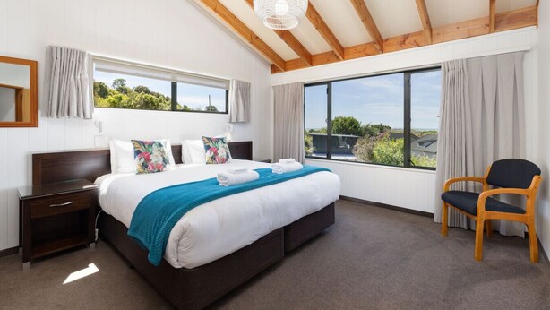 A modern bedroom with a large bed, wooden beams, a view of greenery, and a chair. - Superior Two Bedroom Sea View (Kaiteriteri)