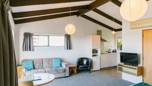 A modern living room with a sofa, a dining table, a kitchen area, and a television. - Studio Sea View Upper Level (Kaiteriteri)