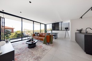 A modern living room with a large window, a sofa, a coffee table, and a dining area. - The Horizon Apartments by Urban Rest (Randwick)