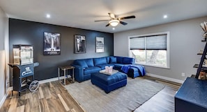 A modern living room with a blue sectional sofa, a small coffee table, a ceiling fan, and a popcorn machine. - Snowbird, ASU, Fall Special @ Lakeview Home with movie theater, pool, firepit (Mesa)