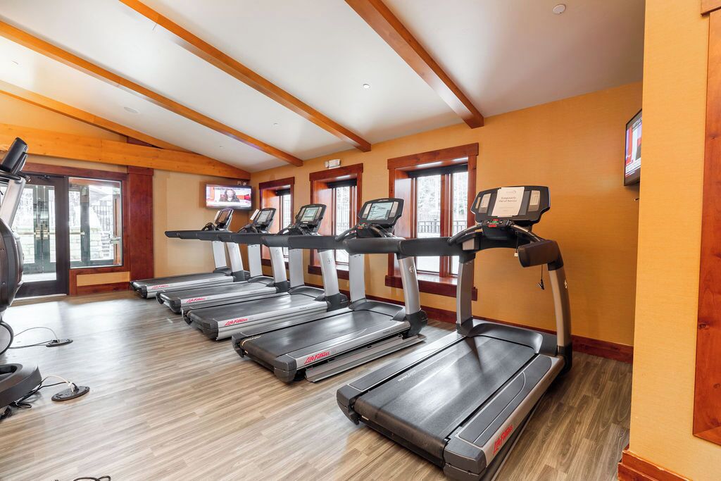 A gym with treadmills and a stationary bike, wooden beams on the ceiling, and large windows.