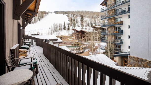 Une terrasse en bois avec des sièges extérieurs, offrant une vue sur une piste de ski enneigée et sur les bâtiments environnants.