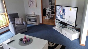 Olohuoneessa on harmaa sohva, valkoinen sohvapöytä, jossa on kaukosäädin, lasijalustalla oleva televisio ja takka.