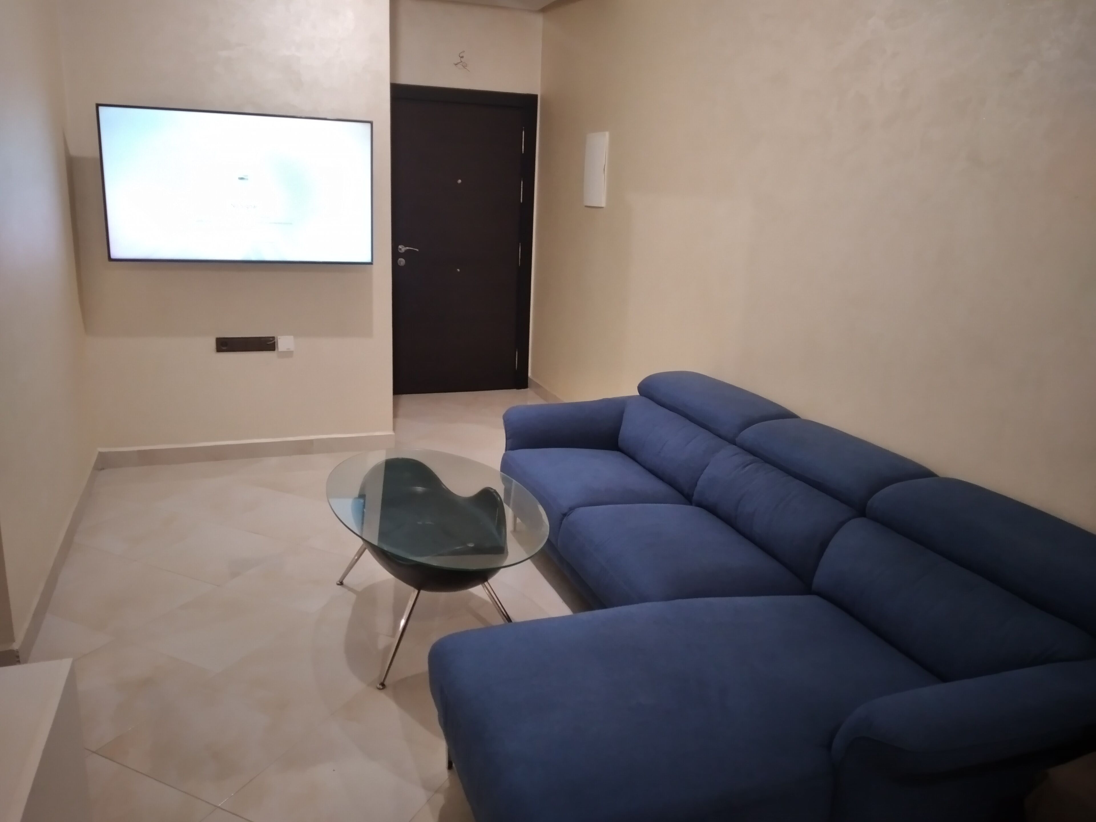 A living room with a blue sofa, a glass coffee table, and a TV mounted on the wall.
