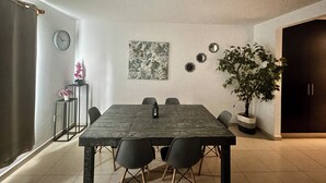 Zona de menjador amb una taula gran, cadires, un rellotge, un quadre floral i una planta en test.