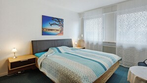 Un dormitorio con cama, mesitas de noche, un velador, una ventana con cortinas y un cuadro colgado en la pared.