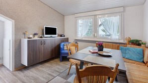Una cocina moderna con televisor integrado, un comedor con mesa y sillas, y una zona de estar acogedora con cojines.