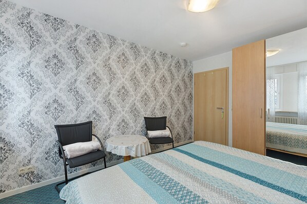 A room with a patterned wallpaper, two chairs, a small round table, a bed with a striped bedspread, and a wooden wardrobe. - Apartment "Haus der Gemütlichkeit" with Balcony & Wi-Fi (Konstanz)