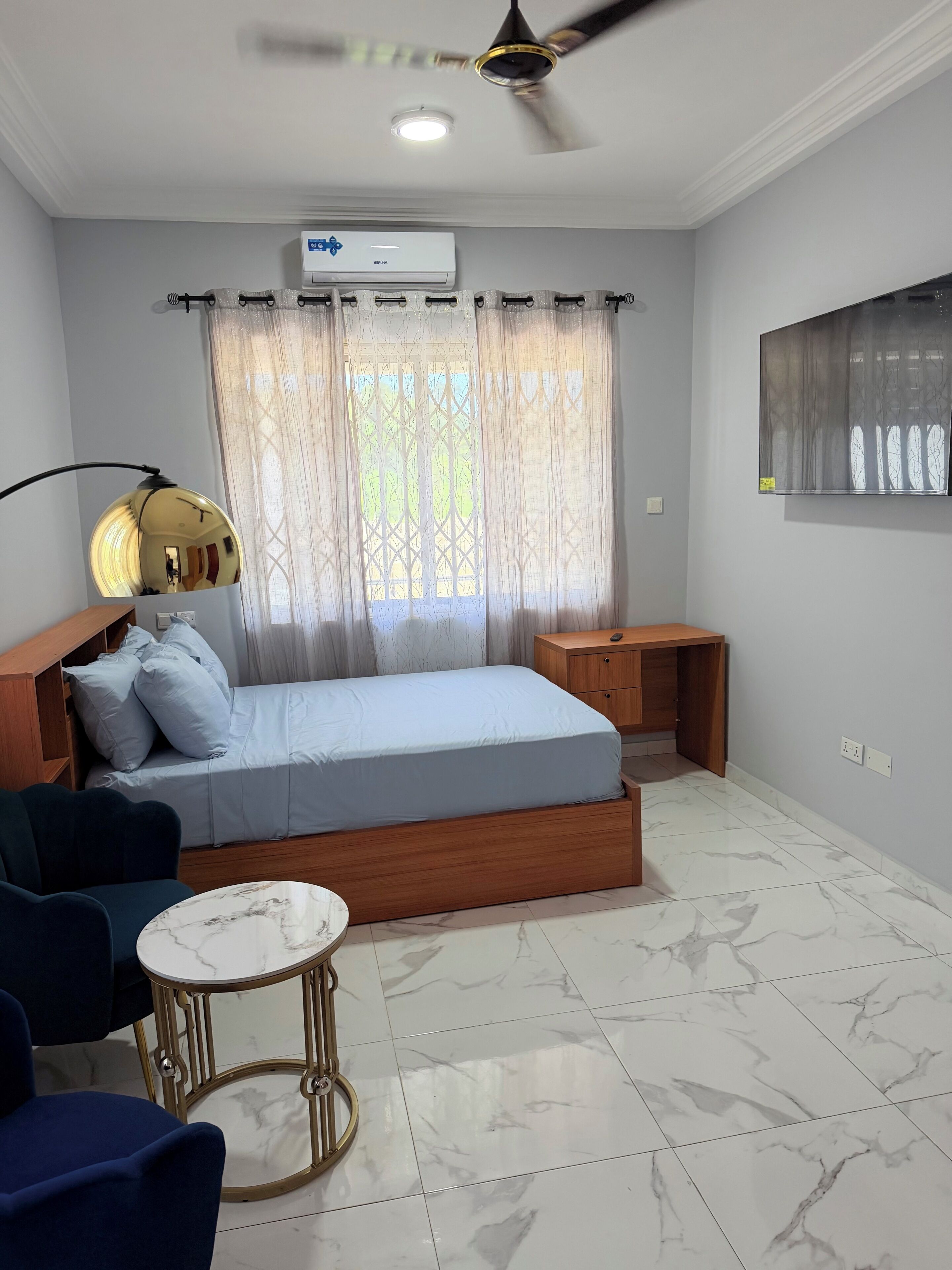 Un dormitorio con cama, mesita de noche, ventilador de techo, unidad de aire acondicionado, ventana con cortinas y una obra de arte colgada en la pared.