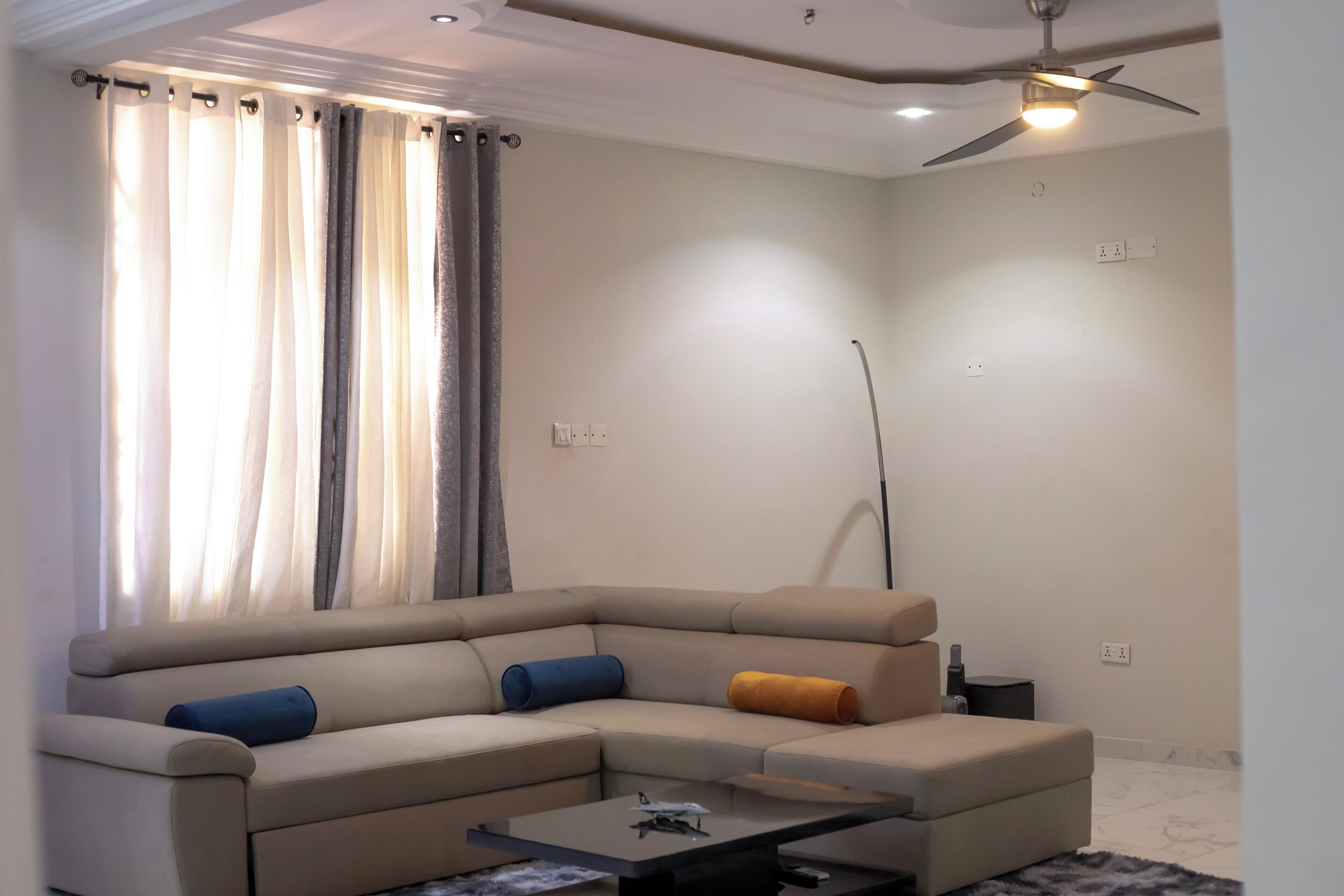 A modern living room with a beige sectional sofa, a glass coffee table, and a ceiling fan.