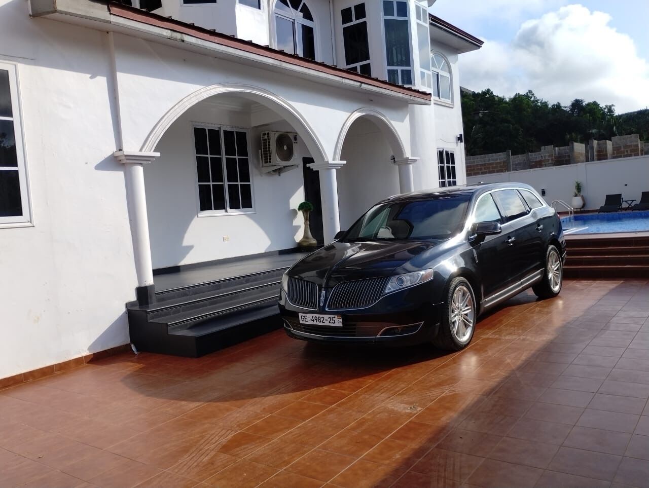 A black car parked in front of a white building with a tiled roof and a swimming pool in the background.