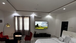 A modern bedroom with a large bed, a flat-screen TV mounted on the wall, a seating area with chairs and a table, and a ceiling with recessed lighting.