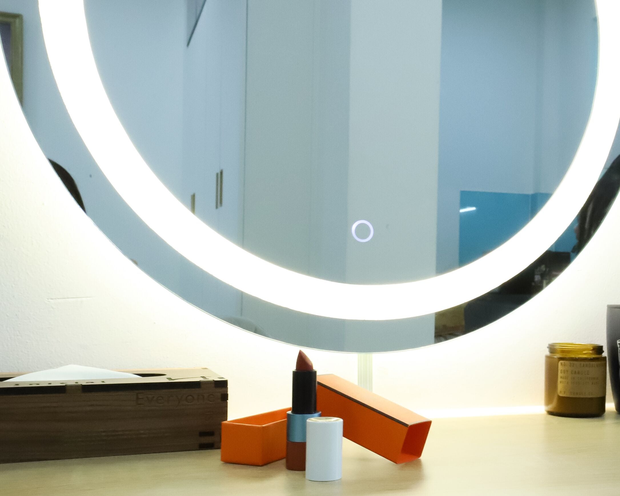 A round mirror with a light, a makeup palette, and a jar of candle wax on a wooden surface.
