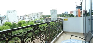 Un balcon orné de balustrades en métal ouvragé, une table en verre et une chaise. En arrière-plan, on aperçoit divers bâtiments et de la verdure.