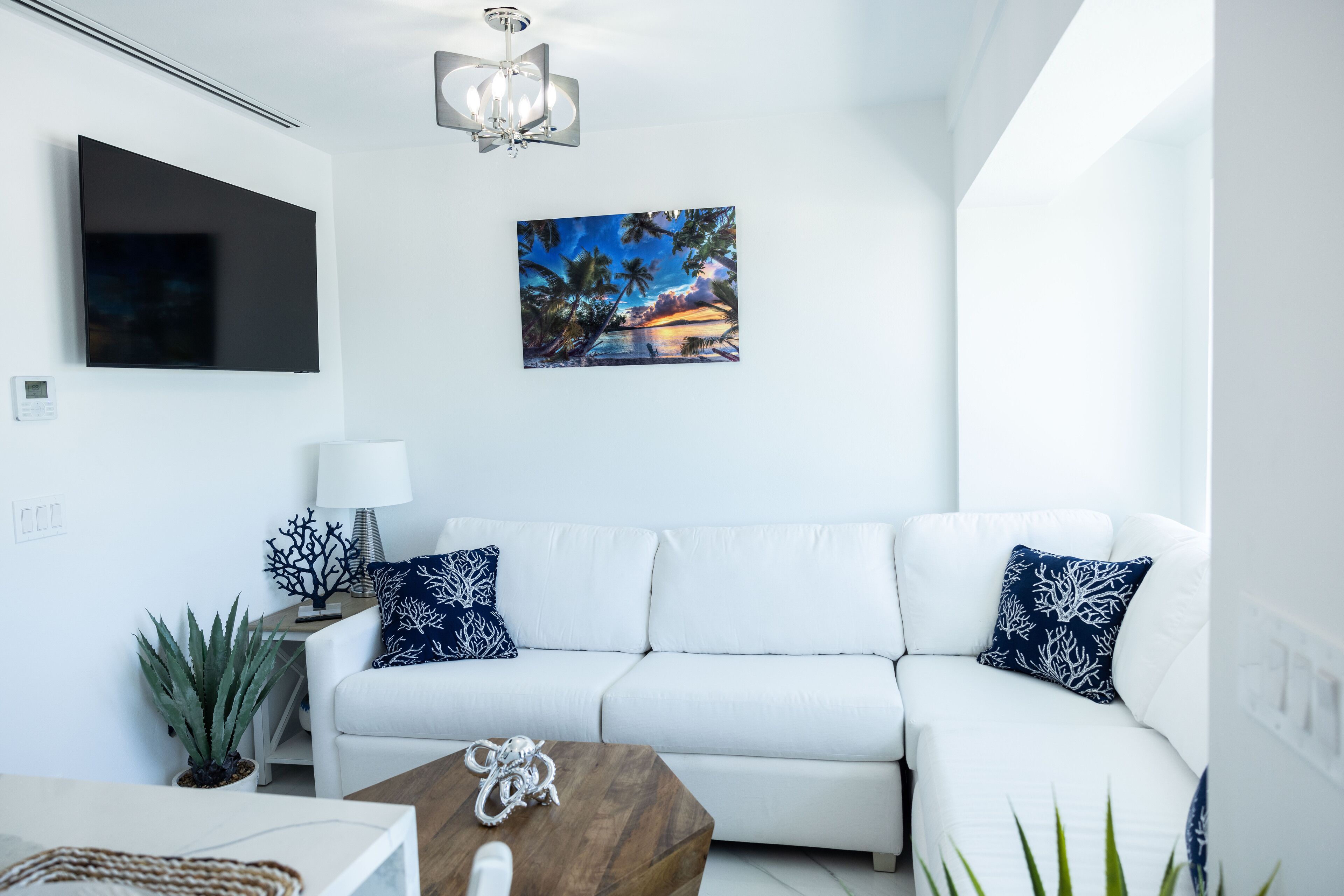 Ein modernes Wohnzimmer mit einem weißen Sofa, einem Holztisch, einem Flachbildfernseher und einem Gemälde, das einen tropischen Sonnenuntergang zeigt.
