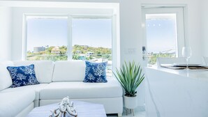 A modern living room with a white sofa, two blue patterned pillows, a white coffee table with a decorative item, a potted plant, and a view of the city through large windows. - Dolphin Suites  (St. John)