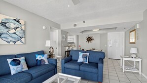 A living room with two blue sofas, a glass coffee table, and a kitchen in the background. - Oceanfront Condominium with Great Amenities (Saint Simons Island)