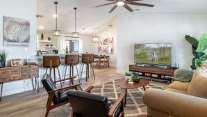 Una sala de estar moderna con televisor grande, asientos cómodos, una zona de cocina y un ventilador de techo.