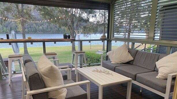 Uma área de estar coberta ao ar livre, com vista para um lago, mobiliada com um sofá, cadeiras e uma mesa.
