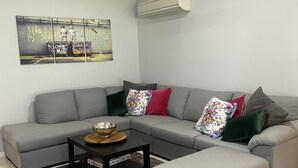 Uma sala de estar moderna com um sofá modular cinza, uma mesa de centro e um banquinho cinza. Há uma pintura na parede e um ar condicionado acima do sofá.