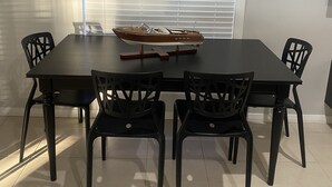 Uma área de refeições com uma mesa retangular e quatro cadeiras, um tabuleiro decorativo sobre a mesa e uma janela com persianas.