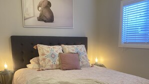 Quarto com cama grande, cabeceira, almofadas decorativas, abajur e quadro na parede.