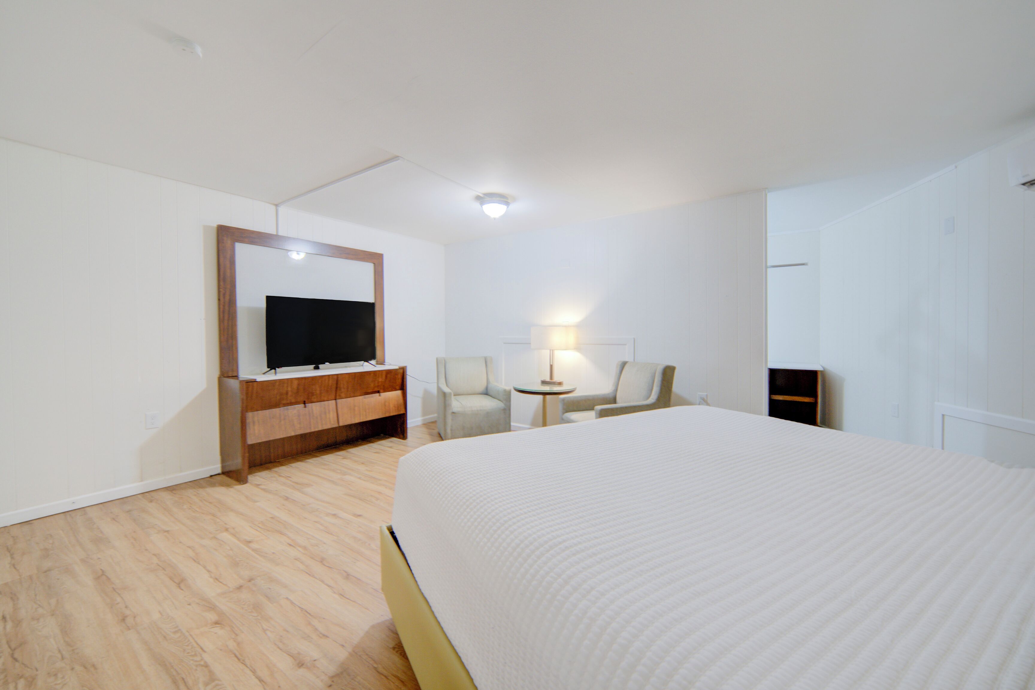 A hotel room with a large bed, a TV on a wooden cabinet, two chairs, a small table with a lamp, and a wardrobe.