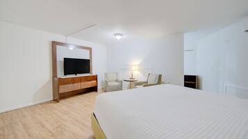 A hotel room with a large bed, a TV on a wooden cabinet, two chairs, a small table with a lamp, and a wardrobe.