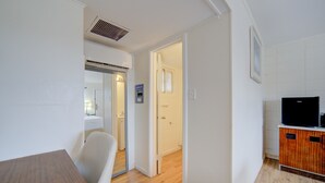 A modern, well-lit room with a wooden dining table, a white chair, and a television on a wooden cabinet. A bathroom with a shower and a toilet is visible through an open door. - Cannon Inn By Capital O Gonzales Tx (Gonzales)
