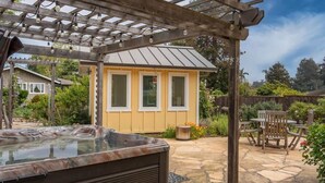 A hot tub under a wooden pergola with a stone patio, surrounded by greenery and a small wooden structure. - Butterfly Beach House (Santa Cruz)