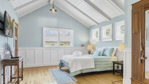 A bedroom with a curved bed, a wooden nightstand, a lamp, and a window with blinds. - Butterfly Beach House (Santa Cruz)