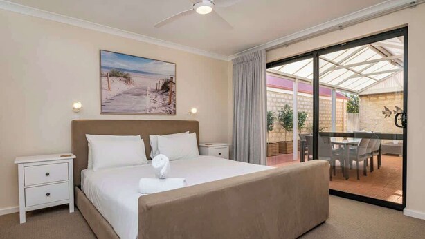 Una camera d'albergo con un letto grande, un comodino, una ventola a soffitto, un quadro appeso al muro e una porta scorrevole in vetro che dà su uno spazio esterno con un tavolo e sedie.