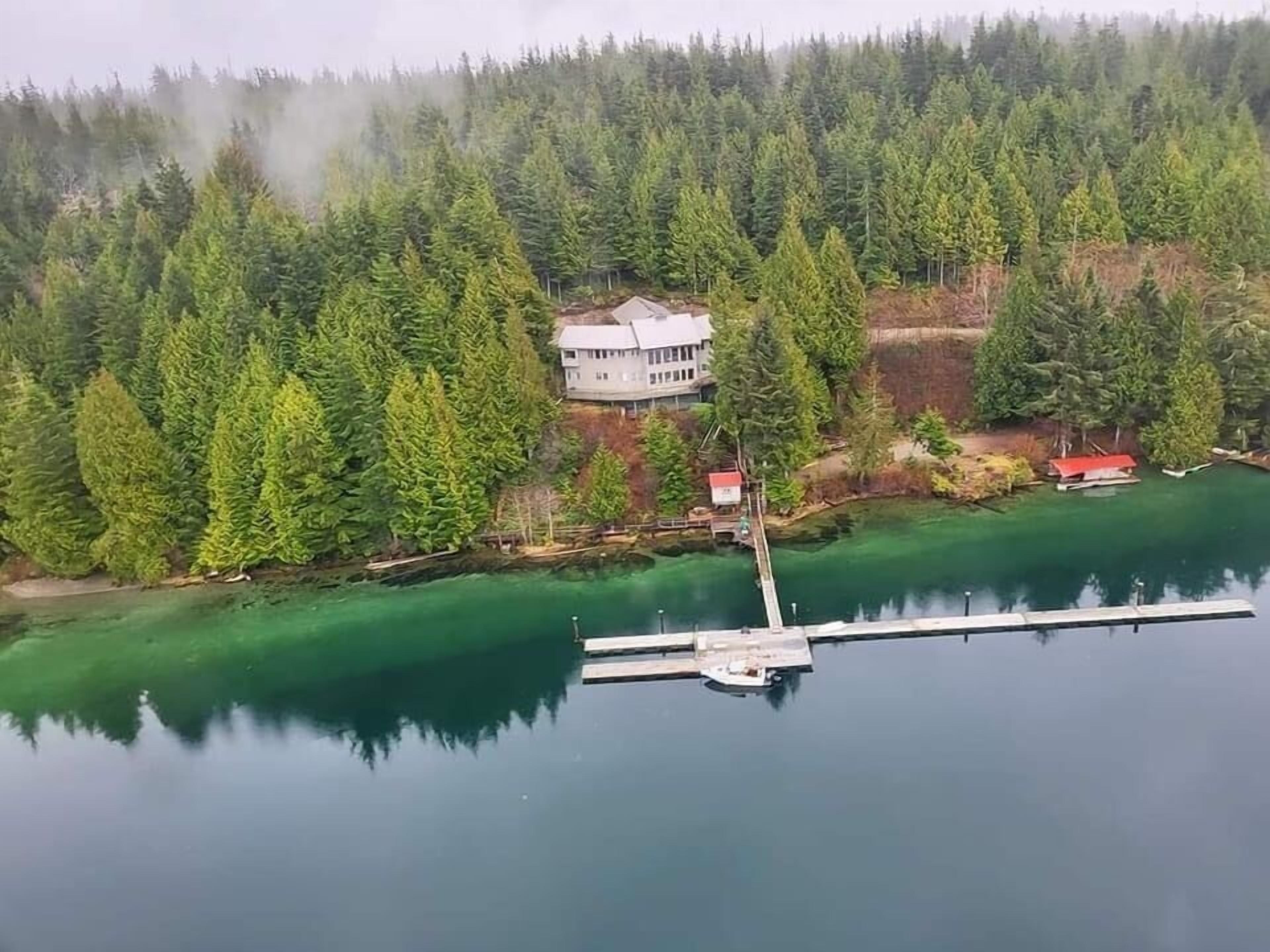 A house surrounded by dense forest, a dock extending into a calm lake, and a small red-roofed structure near the house.