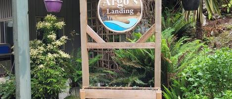 A wooden sign with the text "Argo's Landing" in a circular design, surrounded by lush greenery and hanging planters.
