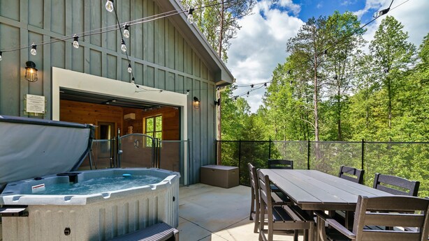A patio with a hot tub, wooden table, and chairs, surrounded by a wooden fence and trees. - Modern cabin w/heated pool & hot tub next to pkwy (Sevierville)